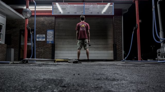 Person stands alone in front of closed car wash station at night. Ideal for themes of loneliness, solitude, or contemplative moments. Can be used in advertisements or media related to urban life, automotive services, or night photography.