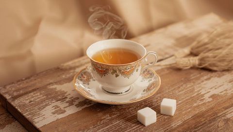 Steaming ornate porcelain teacup on rustic wooden table with sugar cubes and dried wheat