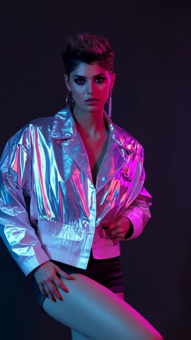 Vertical fashion video showing model adjusting silver jacket under neon pink, cyan lights