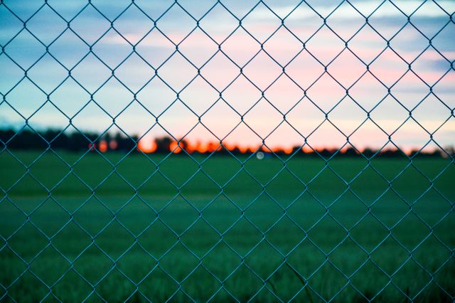 Chain link fence in foreground with soft, vibrant sunset and green field in the background. Useful for illustrating themes of security, barriers, protection, outdoor activities, or rural and urban environments. Suitable for blog posts, articles, marketing materials, or website backgrounds.