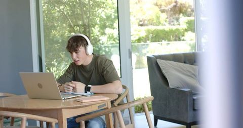 Teenage boy studying at home wearing headphones with laptop by sliding glass doors