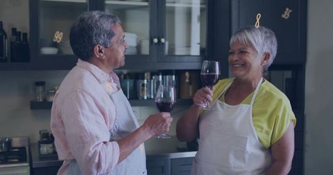 Senior couple toasting with red wine while cooking in cozy home kitchen, wearing aprons
