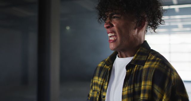 Young man with curly hair and plaid shirt screaming in derelict indoor space, expressing anger and frustration. Useful for depicting intense emotions, mental health issues, or storytelling elements centered around conflict.