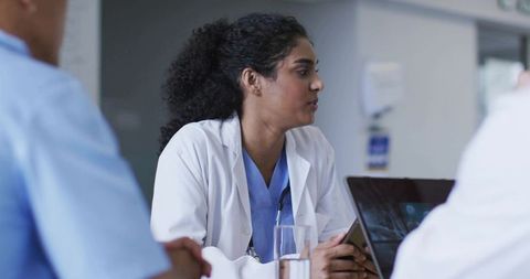 Female physician leading clinical team discussion holding tablet and laptop in hospital meeting