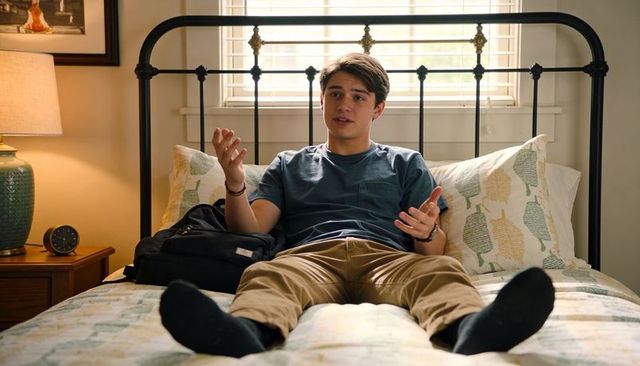 Young student lounging on bed gesturing while talking with backpack and patterned pillows