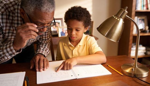 Grandparent tutoring child in math at home desk with lamp and spiral notebook