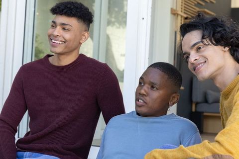 Three diverse teen friends sitting on porch steps laughing and relaxing in knit sweaters, daylight