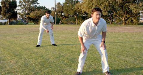 Two Cricketers Crouching in Sunlit Park Practicing Fielding Drills