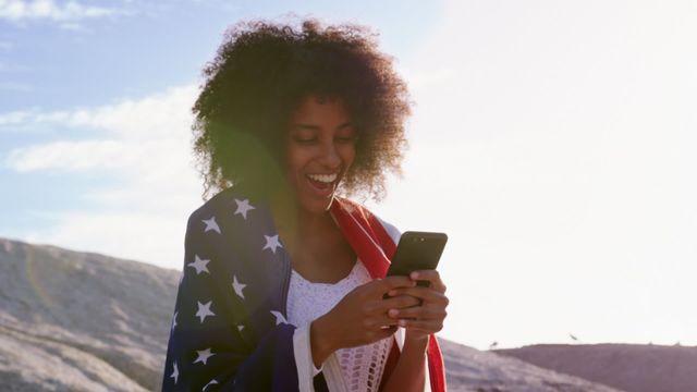 Young African American woman joyfully engaging with her smartphone while wrapped in a vibrant American flag. In a scenic outdoor setting with sunlit hills and a blue sky backdrop, she connects and expresses celebration or patriotism. Ideal for concepts related to national pride, digital communication, or lifestyle imagery emphasizing joy and freedom.