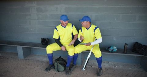 Two diverse baseball teammates sitting in dugout talking and holding bat and duffel bag