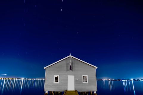 Solitary boathouse at night with star trails and mirror-like harbor reflection