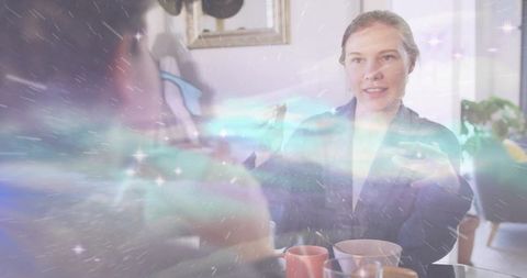 Woman chatting animatedly at cozy dining table with warm pastel light overlay and mugs