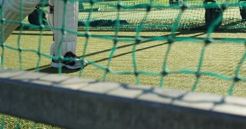 Cricket batter standing in practice net wearing pads and trainers on artificial turf