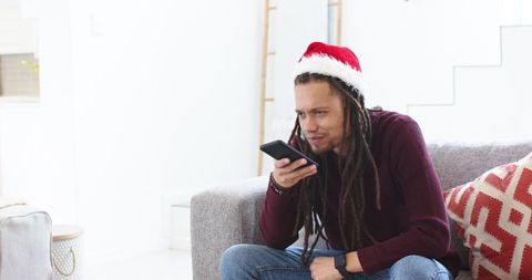 African american man wearing santa hat speaking into phone on sofa for virtual holiday chat