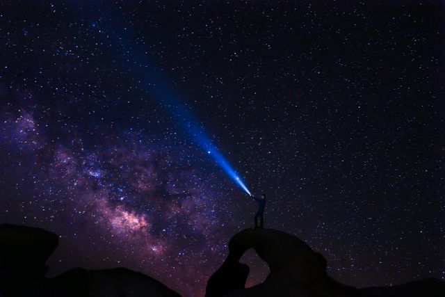 Person standing on rocky arch, shining flashlight into starry night sky, capturing beauty of the Milky Way. Perfect for themes of exploration, night photography, astronomy enthusiasm. Suitable for use in blogs, educational content, travel ads, science and astronomy promotions.