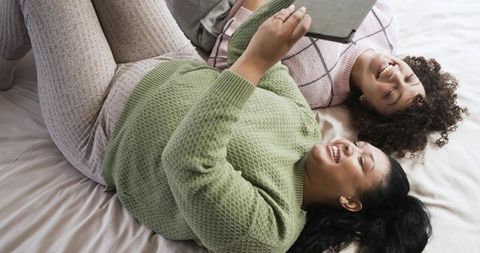 Laughing friends in cozy pajamas sharing tablet and showing ring while relaxing on bed