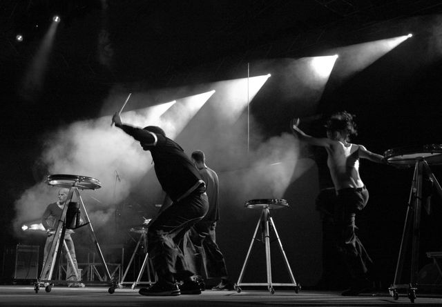 Group of drummers performing on stage with dramatic spotlights in black and white. Ideal for use in music event promotions, concert posters, or blog posts about live performances and music culture.