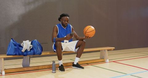 African american basketball player checking phone while spinning ball on gym bench pre-game
