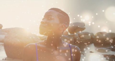 Sunlit black woman standing at rocky shore lifting face to sun in purple swimsuit