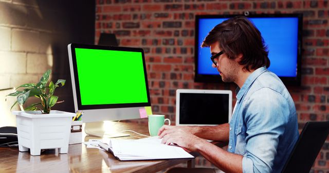 Man working at desk with green screen computer display in modern office with industrial brick walls. Image suitable for technology, business, and productivity concepts. Green screen can be used for customization of the display content.