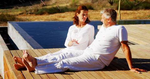 Mature couple relaxing and talking on wooden pool deck in white outfits