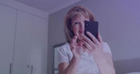 Senior woman holding smartphone smiling while making video call from bedroom mirror