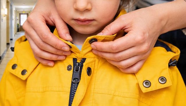 Caregiver fastening yellow raincoat collar for child while preparing for school in hallway