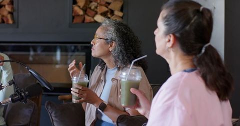 Two women sipping green smoothies recording podcast by fireplace with microphone
