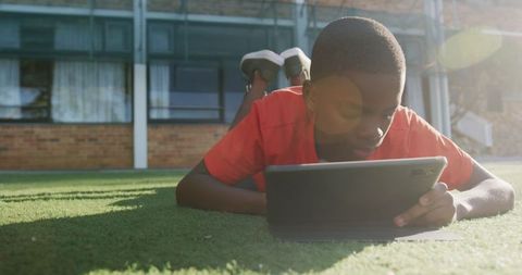 Boy Using Tablet for Study on Grass Outdoors, School in Background