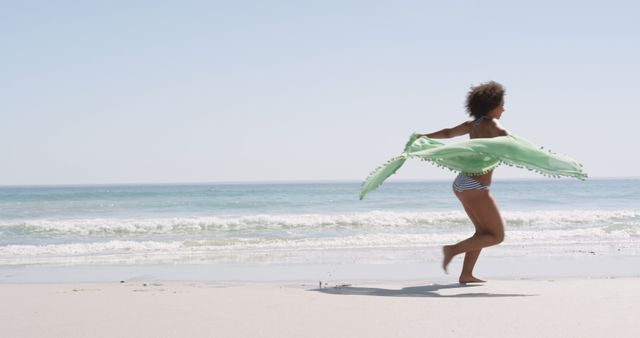 A woman holding and waving a green scarf with tassels is running along the beach on a sunny day. Ideal for use in vacation and travel advertisements, summer clothing promotions, or lifestyle blogs focused on enjoying the beach and outdoor activities.