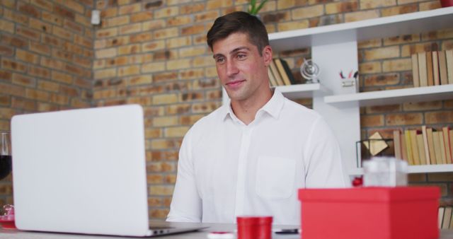 Professional man wearing white shirt using laptop in home office with a glass of wine and gift box in the foreground. Brick wall and bookshelves in the background indicate a cozy working environment. Perfect for content related to remote work, business professionals, productivity, and home office setups.