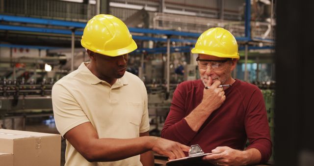 Workers wearing safety helmets and protective gear are discussing production plans on a clipboard in a manufacturing plant. This image can be used to depict industrial environments, teamwork, engineering processes, and safety protocols in various media such as corporate materials, websites, and training manuals.