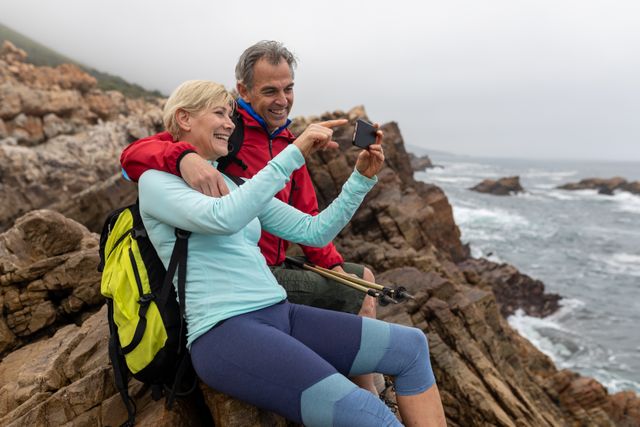 Senior couple enjoying a hiking adventure by the rocky coast, using a smartphone to capture memories. Ideal for promoting active lifestyles, retirement activities, travel destinations, and outdoor adventures. Perfect for use in advertisements, travel blogs, and health and wellness campaigns.