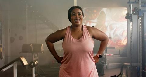 African american woman smiling and posing in gym wearing pink tank and smartwatch
