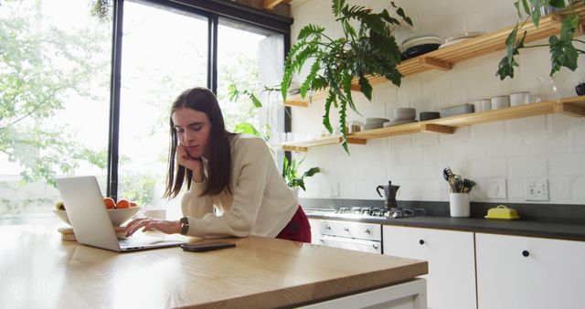 Young woman working on her laptop in a modern kitchen with natural light and open shelving. Ideal for concepts of remote work, home office setups, modern living, and productivity in a casual setting. Can be used for promoting work-from-home lifestyles, interior design ideas, and home office products.
