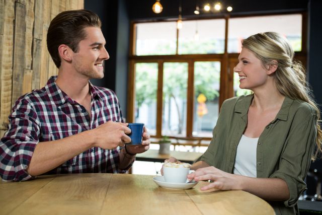 Young couple sitting at a wooden table in a cozy cafe, enjoying coffee and engaging in conversation. Ideal for use in advertisements for coffee shops, cafes, or social media posts promoting relaxation and social interaction.