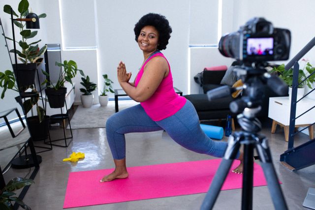 African American woman in sportswear filming her exercise routine on a yoga mat in a living room. She is smiling and performing a yoga pose while being recorded by a camera on a tripod. The room is decorated with plants, creating a fresh and motivating environment. This image is ideal for content related to home workouts, fitness vlogging, online exercise classes, health and wellness during self-isolation, and promoting an active lifestyle during the COVID-19 pandemic.