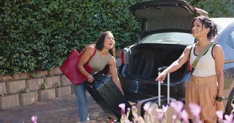 Two friends loading luggage into car on sunlit driveway preparing for road trip