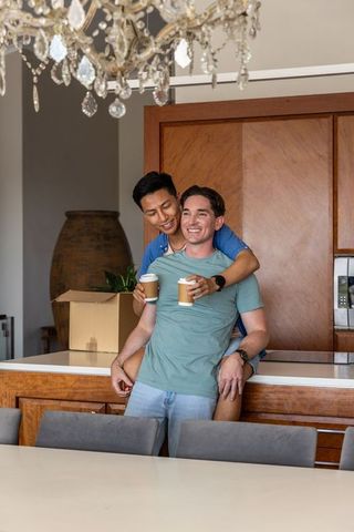 Multicultural gay couple hugging at kitchen island with coffee and moving boxes, new home joy