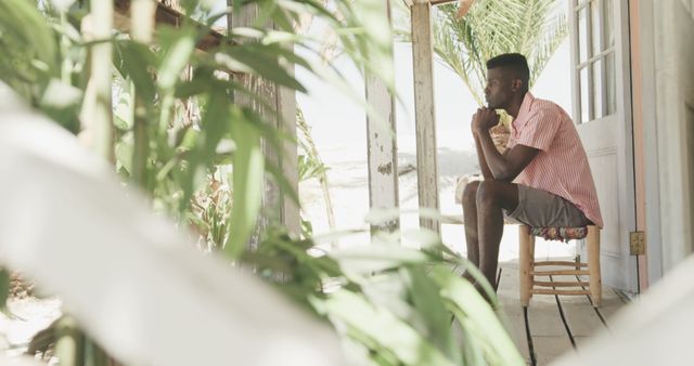 Man is sitting on a porch, surrounded by vibrant greenery, deep in thought. Ideal for themes of relaxation, contemplation, solitude, and connection with nature. Suitable for use in travel advertisements, mental wellness promotions, or lifestyle blogs focused on relaxation and personal time.