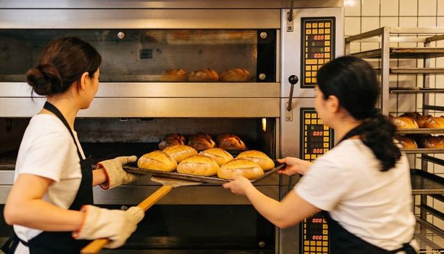 Female bakers removing hot tray from commercial oven in busy artisan bakery production