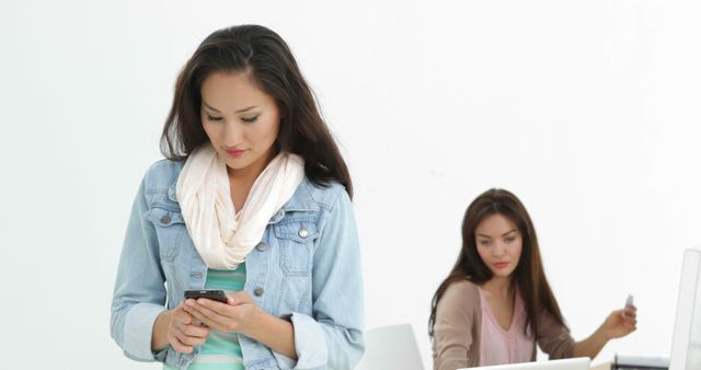 Two women engaging in different activities in a modern office. One woman looks focused on her smartphone, likely checking messages or browsing, while the other woman is working at her computer. This could be used to depict multitasking, modern office environments, professional communication, or digital lifestyles.