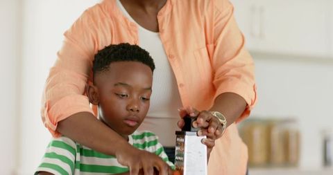 African american mother guiding young son grating carrot at home kitchen counter
