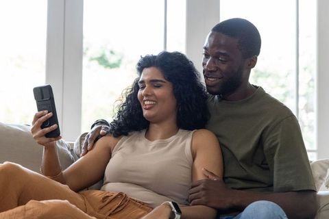Couple relaxing on couch sharing smartphone, indian woman and african american man