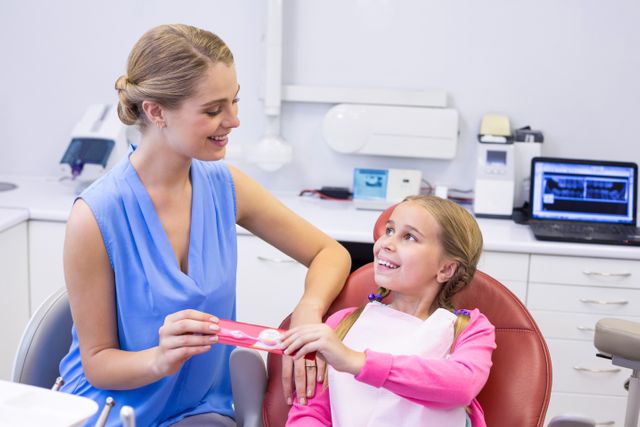 Mother giving toothbrush to daughter at dental clinic. Both are smiling, promoting positive dental care experience. Ideal for use in dental care advertisements, pediatric dentistry promotions, family healthcare brochures, and oral hygiene educational materials.