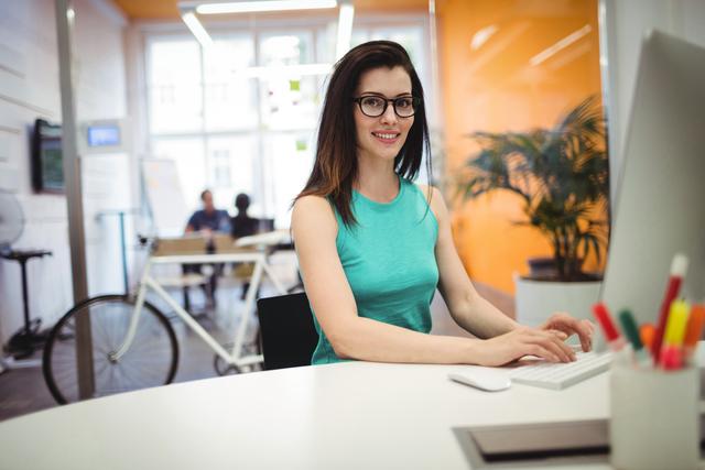 Confident businesswoman with glasses typing at her computer in a bright, modern office. Ideal for use in business, professional, and workplace themes to illustrate productivity, professionalism, and modern office environments.