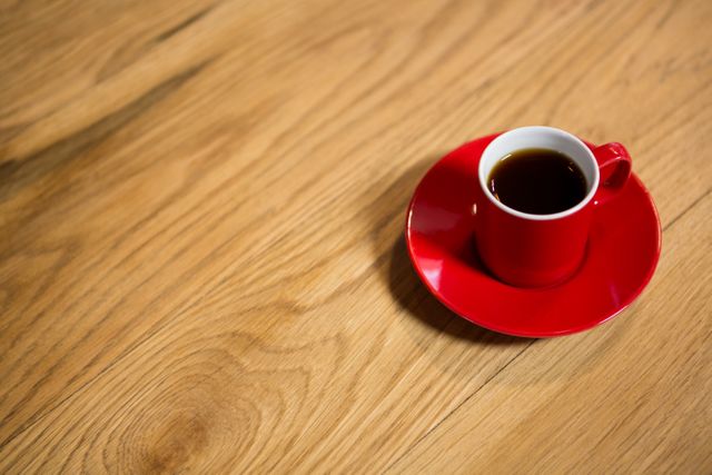 Red coffee cup with saucer on wooden table, perfect for illustrating coffee culture, cafe ambiance, or morning routines. Ideal for use in blogs, social media posts, advertisements, and websites related to coffee shops, relaxation, and lifestyle.