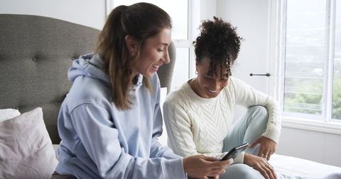 Two women sharing smartphone on bed in bright bedroom smiling, wearing hoodie and sweater