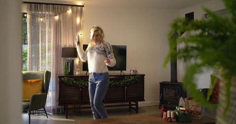 Cozy holiday living room woman dancing with smartphone, lights, garland and wrapped gifts