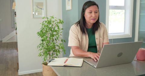 Mature asian woman working from home on laptop at sunlit kitchen counter
